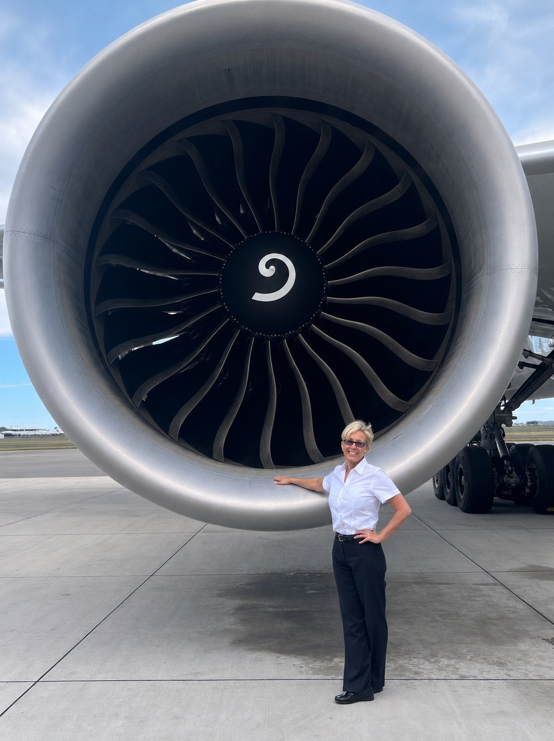 Micki in a white shirt and slacks beside an airliner engine many times her height.